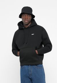 Hanorac negru cu glugă, cu un buzunar frontal, având un logo alb "OBEY". Asortat cu blugi de denim albaștri și o pălărie bucket neagră.