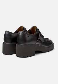 Chaussures en cuir noir avec une semelle plateforme robuste, dotées d'un détail de boucle et d'un dessus texturé. La vue arrière montre le logo et des accents de couture.