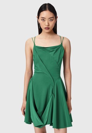 Femme portant une robe verte sans manches avec de fines bretelles et un drapé sur la jupe, debout devant un fond clair uni.
