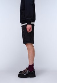 Vista laterale di una persona che indossa pantaloncini cargo neri, scarpe da trekking nere con lacci rossi, calzini viola e una felpa scura su uno sfondo semplice.