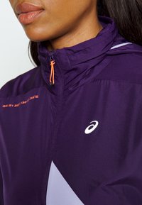 Chaqueta deportiva morada con tirador de cremallera naranja, acentos reflectantes y logotipo blanco. Presenta un diseño geométrico y una textura ligera.