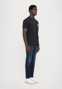 Svart T-skjorte med korte ermer og logo på brystet, kombinert med mørkeblå jeans og hvite joggesko, vist på en stående person.