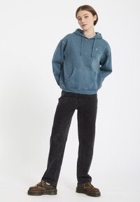 Hoodie in vervaagd zeegroen katoen, met een voorknoopzak. Gecombineerd met losse zwarte corduroy broeken en bruine veterschoenen.