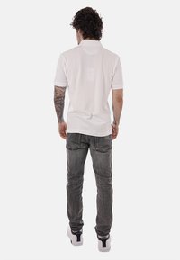 Homme aux cheveux bouclés et tatouages sur les bras, vu de dos, portant un polo blanc, un jean gris et des baskets blanches.