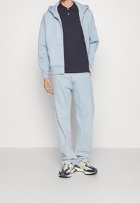 Haut de jogging zippé bleu clair, polo navy, jeans clairs et baskets à motifs. Tissu doux et coupe décontractée pour le confort et une tenue décontractée.