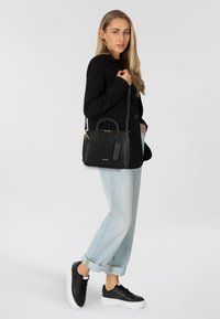 Borsa a mano in pelle nera con superficie texturizzata, manici rotondi e hardware dorato. Indossata con jeans azzurri a maniche rimboccate e sneakers nere.