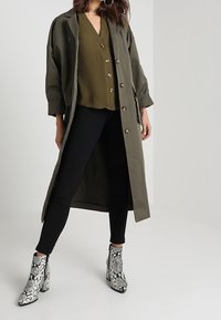 Olivegrüner langer Mantel, über einem dunkelgrünen Oberteil getragen, kombiniert mit schwarzen engen Hosen und ankle boots mit schwarz-weißem Schlangenprint.