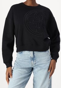 Sudadera negra con logotipo texturizado en la parte delantera, escote redondo y puños acanalados. Combinada con jeans de talle alto en azul claro.