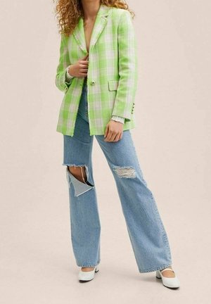 Woman wears light green and white checkered blazer, ripped wide-leg blue jeans, and white shoes, standing on neutral background.