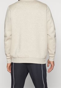 Person som bär en beige långärmad sweatshirt och svarta byxor med vita sidostrickor, sedd bakifrån.