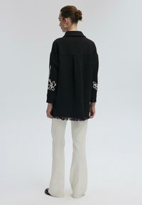 Camicia oversize in misto lana nera con orlo frangiato, caratterizzata da ricami floreali bianchi sulle maniche, abbinata a pantaloni bianchi a zampa.