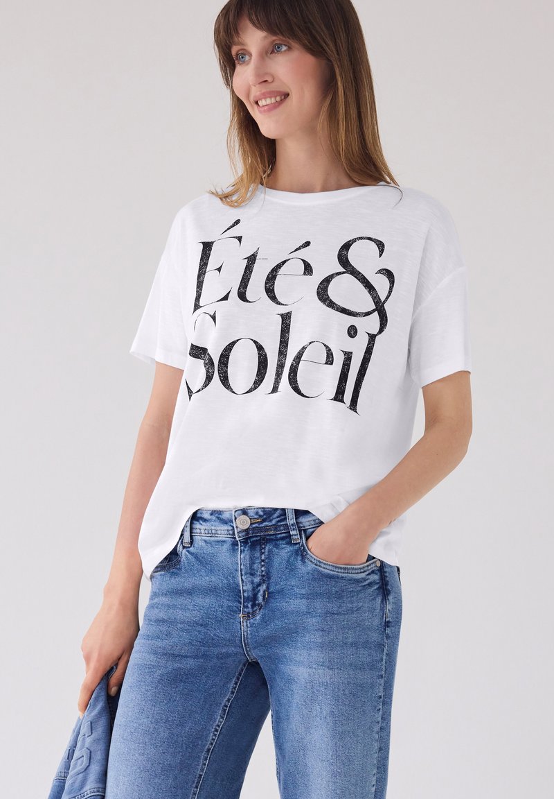 Femme souriante portant un t-shirt blanc avec le texte « Été & Soleil », un jean bleu, tenant un tissu bleu plié dans une main.