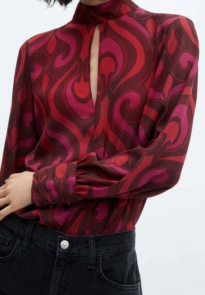 Femme portant un chemisier à manches longues et col montant avec des tourbillons abstraits rouges et violets et un jean noir taille haute.