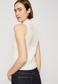 Mouwloze crèmekleurige top met een gestructureerde rug van een patroon, voorzien van een hoge kraag en een slim fit ontwerp. Gecombineerd met donkere spijkerbroeken.