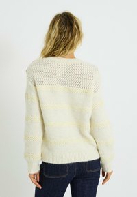Pull en tricot de couleur crème avec des rayures jaunes, présentant un dos texturé et des poignets côtelés. Tissu doux avec une coupe ample et un col rond.