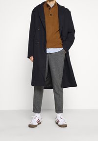 Marineblauwe double-breasted jas over een bruine trui, grijze geruite broek en witte sneakers met rode strepen. Lagen zorgen voor contrast en textuur.