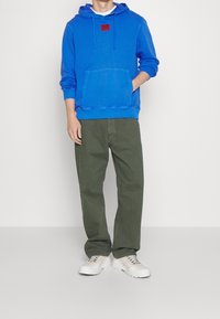Sudadera de algodón azul con bolsillo frontal y etiqueta de la marca, combinada con pantalones de color verde oliva. Zapatillas blancas con cordones completan el atuendo.