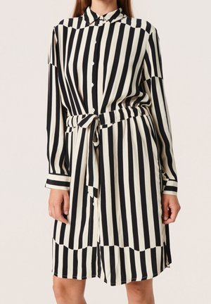 Kobieta ubrana w czarno-białą pionowo prążkowaną sukienkę na guziki z wiązanym paskiem w talii i długimi rękawami.