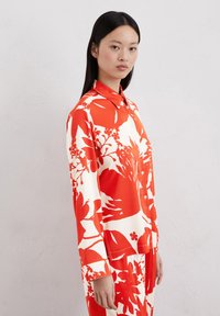 Chemise à boutons rouge et blanche à fleurs avec des manches longues, dotée d'un col et de grands motifs de feuilles et de fleurs dans un tissu lisse.