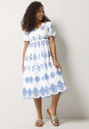 Donna con capelli ricci che indossa un vestito midi bianco con ricamo floreale blu e orlo smerlato, abbinato a sandali intrecciati beige.