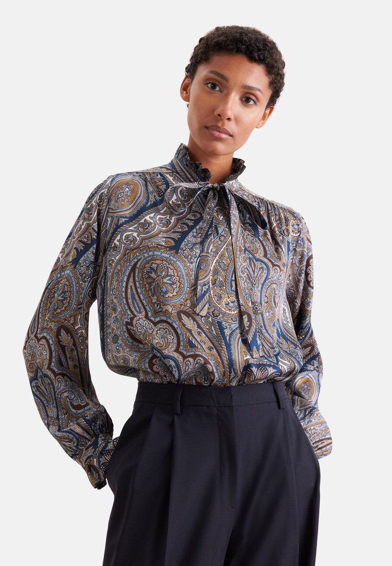 Femme portant un chemisier à col haut à motif cachemire avec des manches longues, rentré dans un pantalon sombre taille haute sur un fond uni.