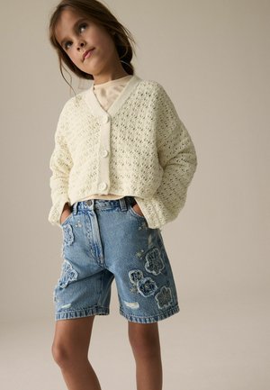 Niña joven con cárdigan de punto color crema y pantalones cortos vaqueros azules con bordado floral, de pie con las manos en los bolsillos.