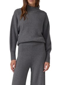 Pull gris en maille avec un col haut, des manches longues et des poignets côtelés. Le tissu semble doux et texturé, offrant une coupe décontractée.