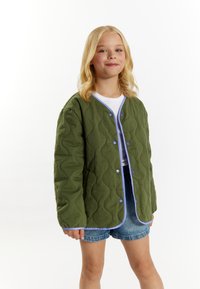 Quilted grøn jakke med rund krave, blå kant og trykknapper. Bæret over en hvid skjorte og kombineret med denimshorts.