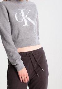 Šedá zkrácená mikina s výrazným bílým logem "CK" a textem "Calvin Klein Jeans". Kombinováno s tmavými tepláky a detaily šňůrky.