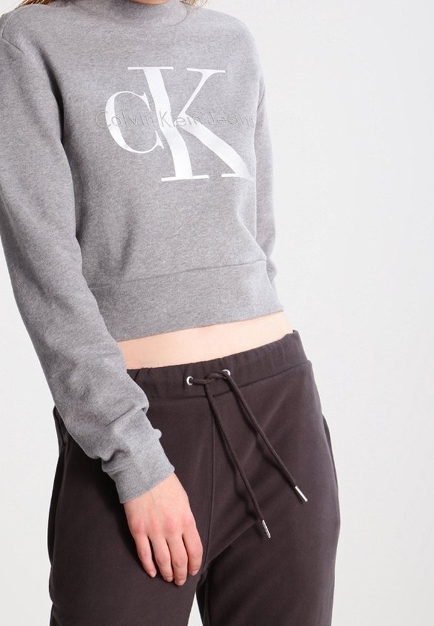 Šedá zkrácená mikina s výrazným bílým logem "CK" a textem "Calvin Klein Jeans". Kombinováno s tmavými tepláky a detaily šňůrky.