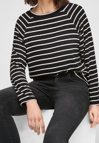 Gestreiftes Langarmshirt in Schwarz und Weiß mit Rundhalsausschnitt, lockerer Passform und gerippten Bündchen. Kombiniert mit schwarzen Jeans.