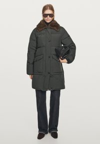 Woman wearing dark green padded coat with brown fur collar, black pants, brown shoes, dark sunglasses, holding black handbag under arm.