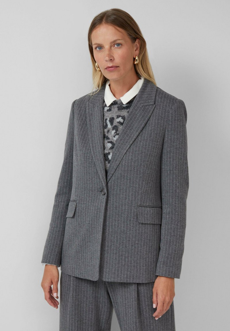 Femme portant un costume à rayures grises avec un pull en cuir de léopard et une chemise blanche à col, se tenant devant un fond clair.