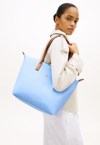 Γυναίκα που φοράει μπεζ μπουφάν και λευκό παντελόνι, κρατώντας μια μεγάλη, ανοιχτό γαλάζια τσάντα ώμου με καφέ χερούλια.