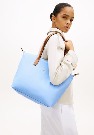 Mujer con chaqueta beige y pantalones blancos llevando un gran bolso tote azul claro con asas marrones sobre el hombro.