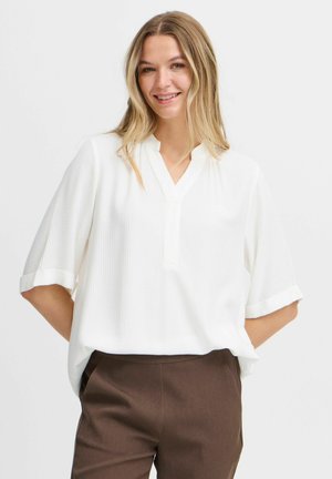 Blouse blanche à manches courtes avec un col à cran, en tissu texturé et avec des poignets retroussés, associée à un pantalon tailleur marron.