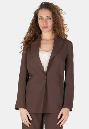 Donna con lunghi capelli ricci che indossa un blazer marrone su misura e pantaloni abbinati sopra una maglietta bianca, in piedi davanti a uno sfondo bianco.