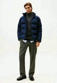 Navy puffer jacket with quilted sections, high collar, paired with a gray cable-knit sweater and gray patterned trousers. Glossy black shoes.