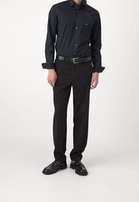 Camicia nera con bottoni, dotata di un logo sottile, abbinata a pantaloni scuri e scarpe nere lucide. Il tessuto sembra liscio con una vestibilità sartoriale.