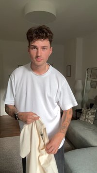 T-shirt oversized branco feito de algodão, posicionado sobre um tecido bege claro. O fundo apresenta um sofá cinza texturizado e elementos decorativos.