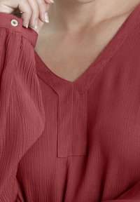 Blouse en tissu rouge texturé avec un col en V, manches froncées et détail de boutons sur l'épaule ; présente un motif de plis subtil.