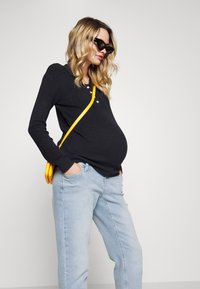 Top de manga longa preto canelado com três botões, jeans azul claro e uma mala tiracolo amarela. Os acessórios incluem óculos de sol pretos.