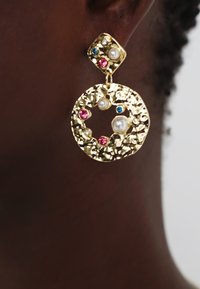 Gullfargede øreringer med teksturert sirkulært design, prydet med perler og fargede edelstener (rosa og blå) på en kvadratisk stud-bunn.