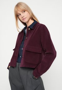Giacca in velluto a coste color bordeaux con collo a revers, chiusura frontale con bottoni e due tasche applicate, indossata sopra una camicia di jeans.