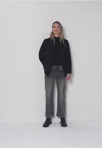 Mujer con cabello rubio, vestida con una chaqueta negra, una blusa negra, jeans de pierna ancha grises y botas negras, de pie frente a una pared lisa de color gris claro.