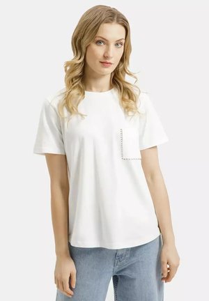 Młoda kobieta o długich blond włosach, ubrana w białą koszulkę z ozdobną kieszonką i jasnoniebieskie dżinsy, stojąca na tle jednolitego tła.