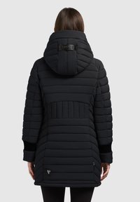 Kobieta w czarnej pikowanej zimowej kurtce z kapturem, widziana od tyłu na jednolitym jasnoszarym tle.