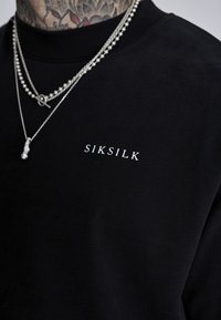 Sudadera de algodón negra con el logo blanco de "SIKSILK" en el pecho. Cadenas de plata superpuestas añaden detalle. Vista de cerca de la parte superior del torso.