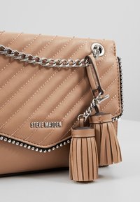 Beige päällystetty nahkakäsilaukku, jossa on hopeinen ketjuhihna, tupsuyksityiskohtia ja niittireunus.