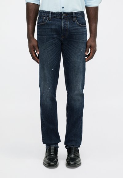 Mann iført mørkeblå, smale jeans med subtile slitasjemerker, hvit skjorte og svarte skinnsko, stående med armene avslappet.